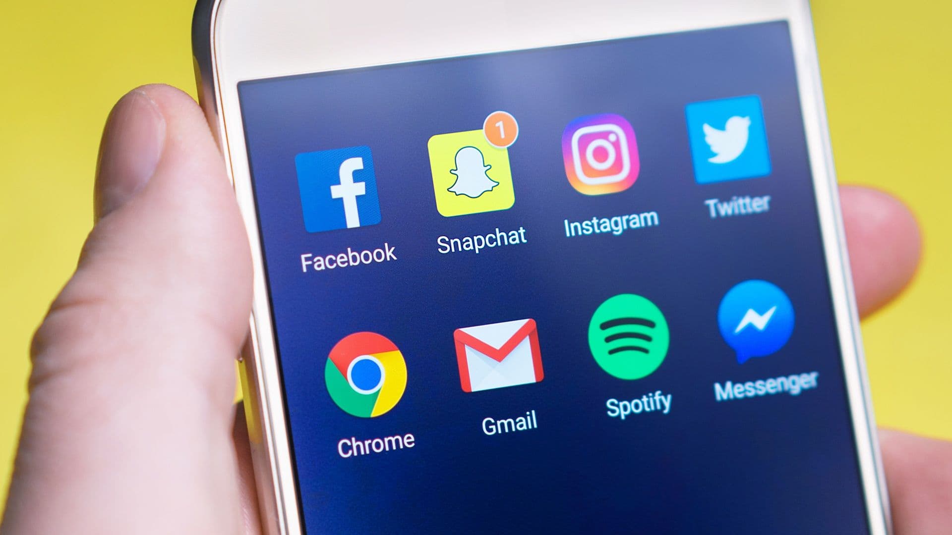 A hand holding a smartphone showing social media app icons including Facebook, Snapchat, Instagram, and Twitter