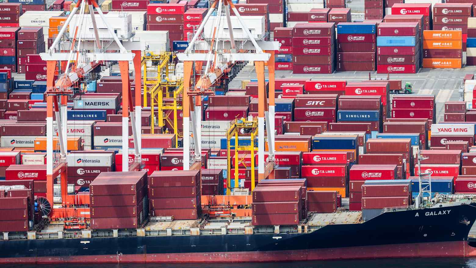 Shipping containers stacked at a port with gantry cranes loading a cargo vessel