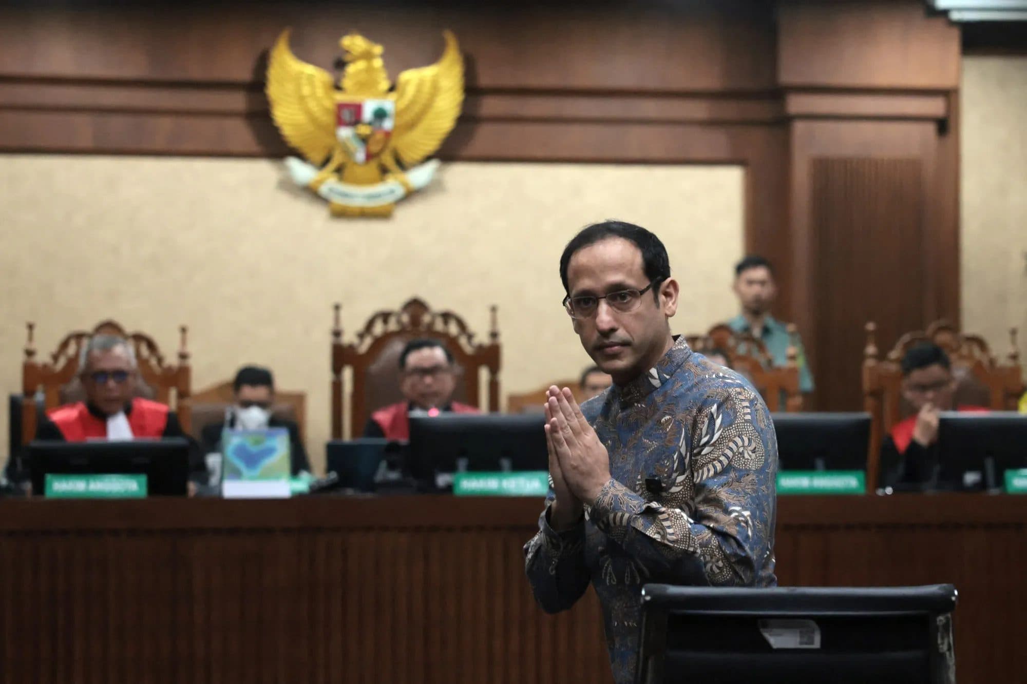 Nadiem Makarim stands with his hands pressed together before judges in a Jakarta courtroom