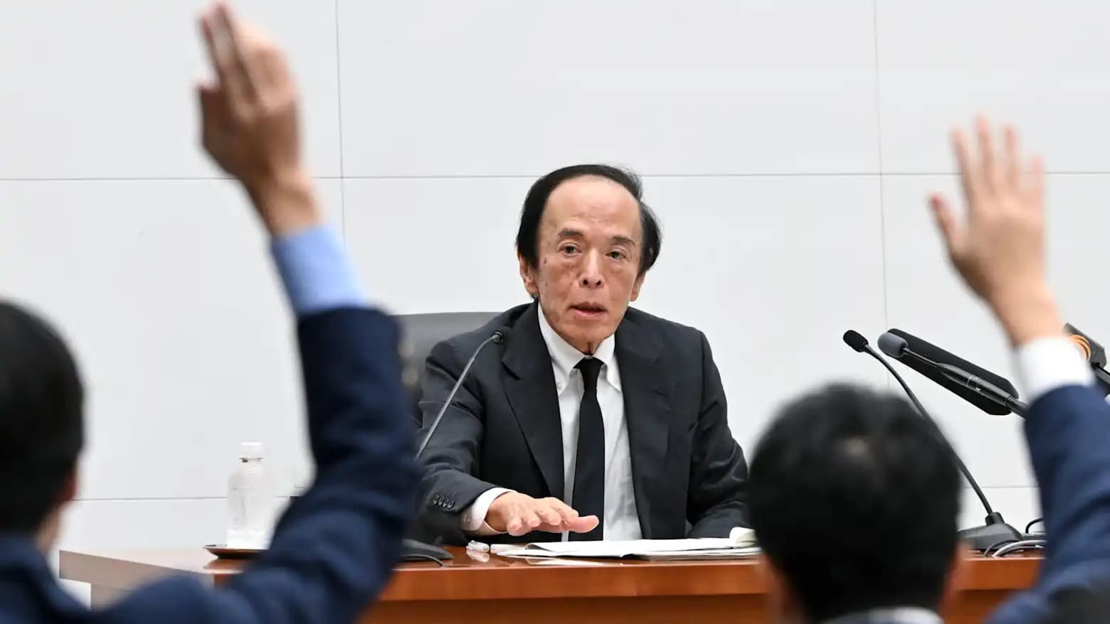 Bank of Japan Governor Kazuo Ueda addressing reporters with hands raised in the foreground