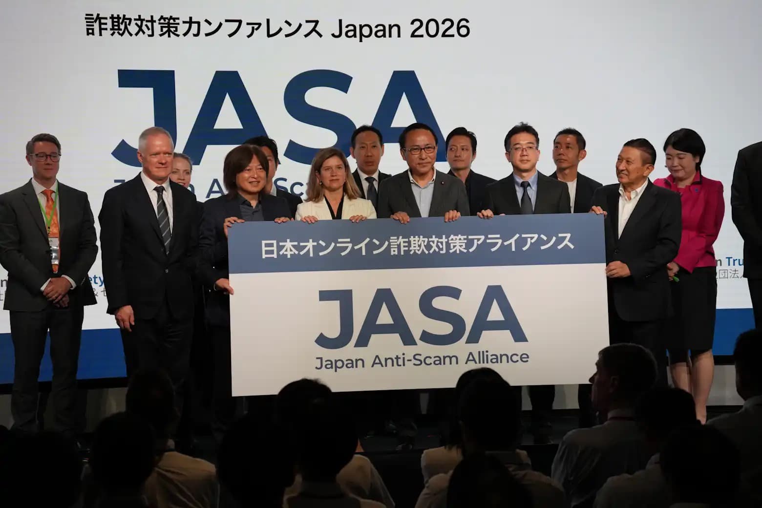 Executives and officials on stage holding a Japan Anti-Scam Alliance (JASA) banner at the Anti-Scam Conference Japan 2026