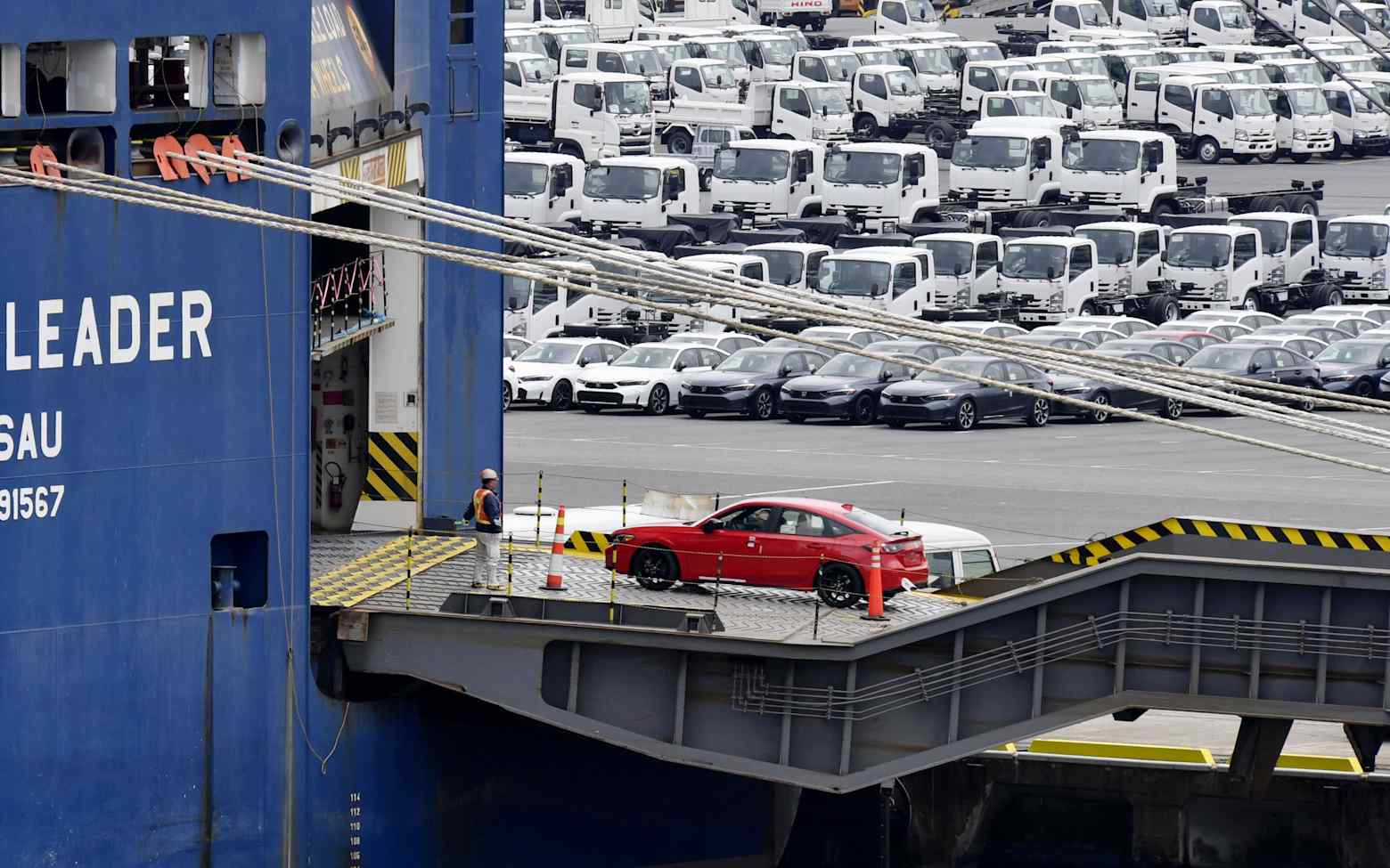 A car is driven onto a vehicle carrier ship for export at a Japanese port, with rows of cars and trucks waiting on the dock
