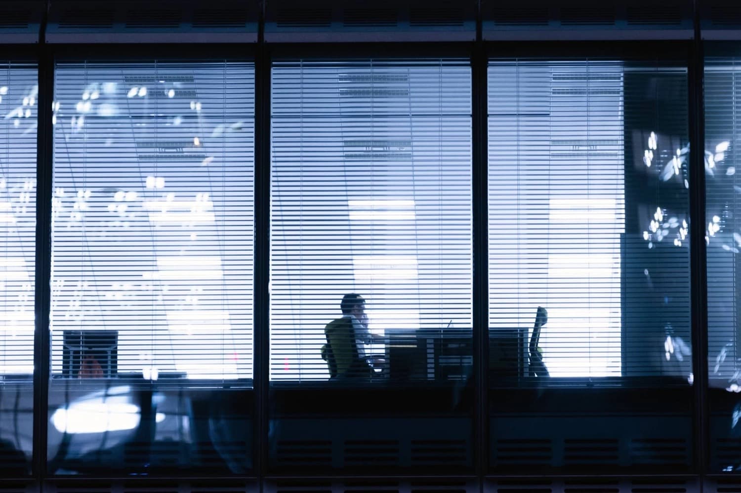 A lone office worker silhouetted at a desk behind window blinds late at night